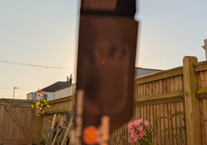 Eclipse glasses held infront of the camera, far enough away to just cover the sun. Behind it a wooden fence and some flowers